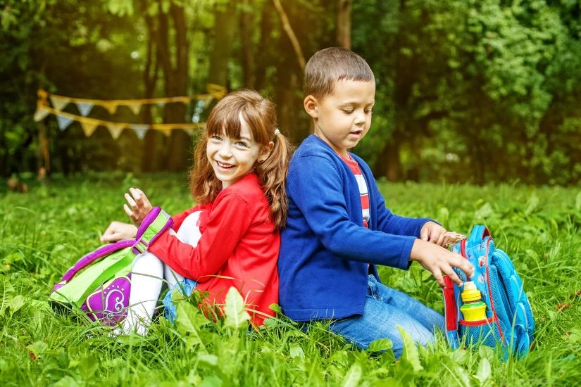 Dečak i devojčica sede na travi i oboje nose ranac na leđima.