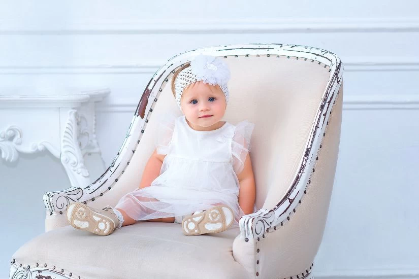 Beba u beloj haljini sedi na stolici.